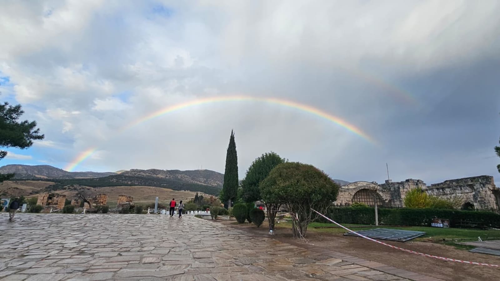 A rainbow over a stone-paved path in Hierapolis, the Roman spa city above the Pamukkale terraces, with a few visitors walking in the distance