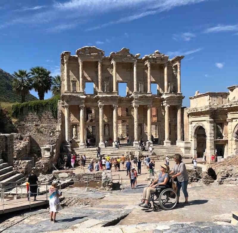 A visitor in a wheelchair with a companion in front of the Library of Celsus at Ephesus