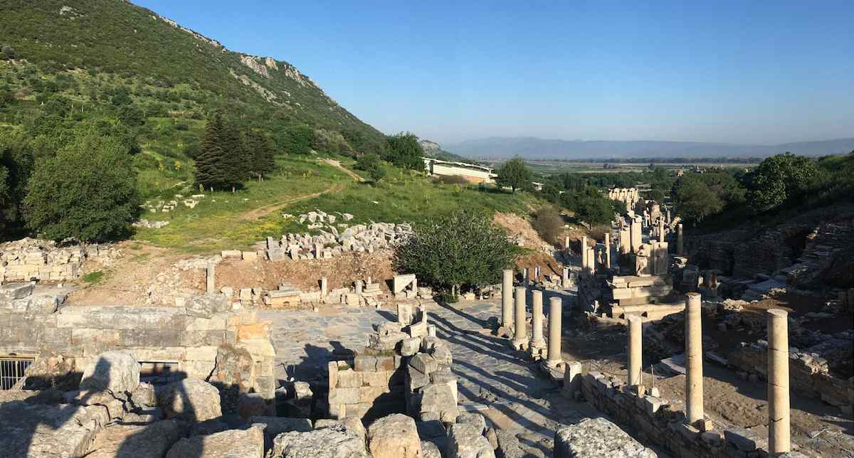 Curetes Street at Ephesus seen from above in early-morning light with no one on it, running down past columns toward the valley, with the white roof of the Terrace Houses on the green hillside