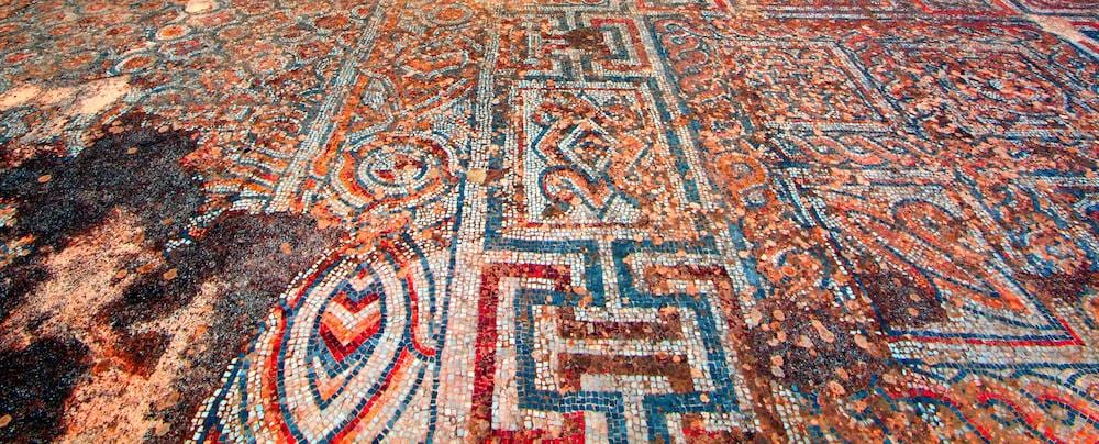 Ancient geometric floor mosaic in red, cream and blue with meander and chevron patterns, laid in small stones.