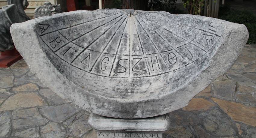 Semicircular ancient marble sundial carved with Greek letters and hour lines, displayed outdoors on a stone plinth.