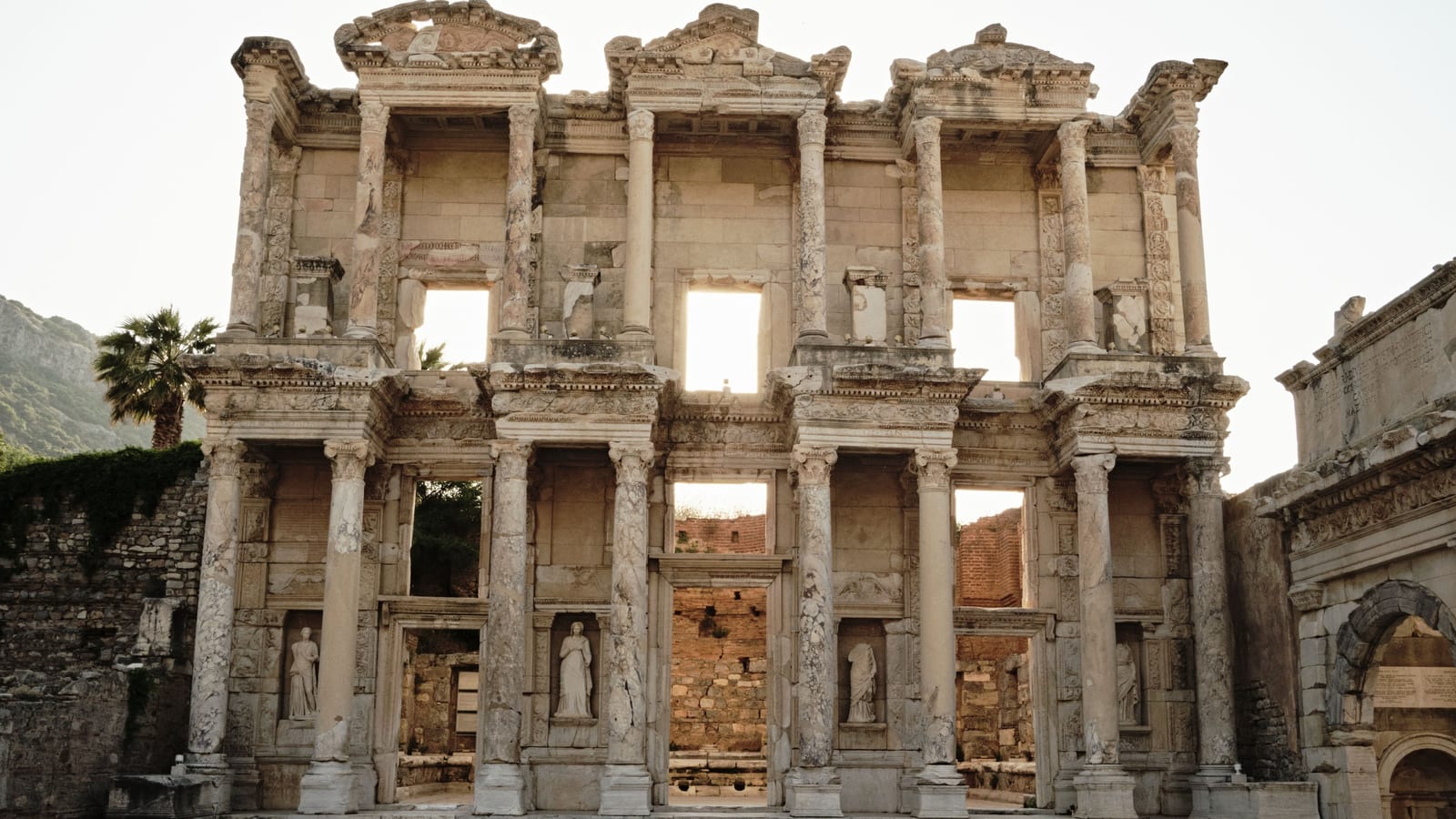 The empty facade of the Library of Celsus at Ephesus in soft early light, with the Gate of Mazaeus and Mithridates at the right