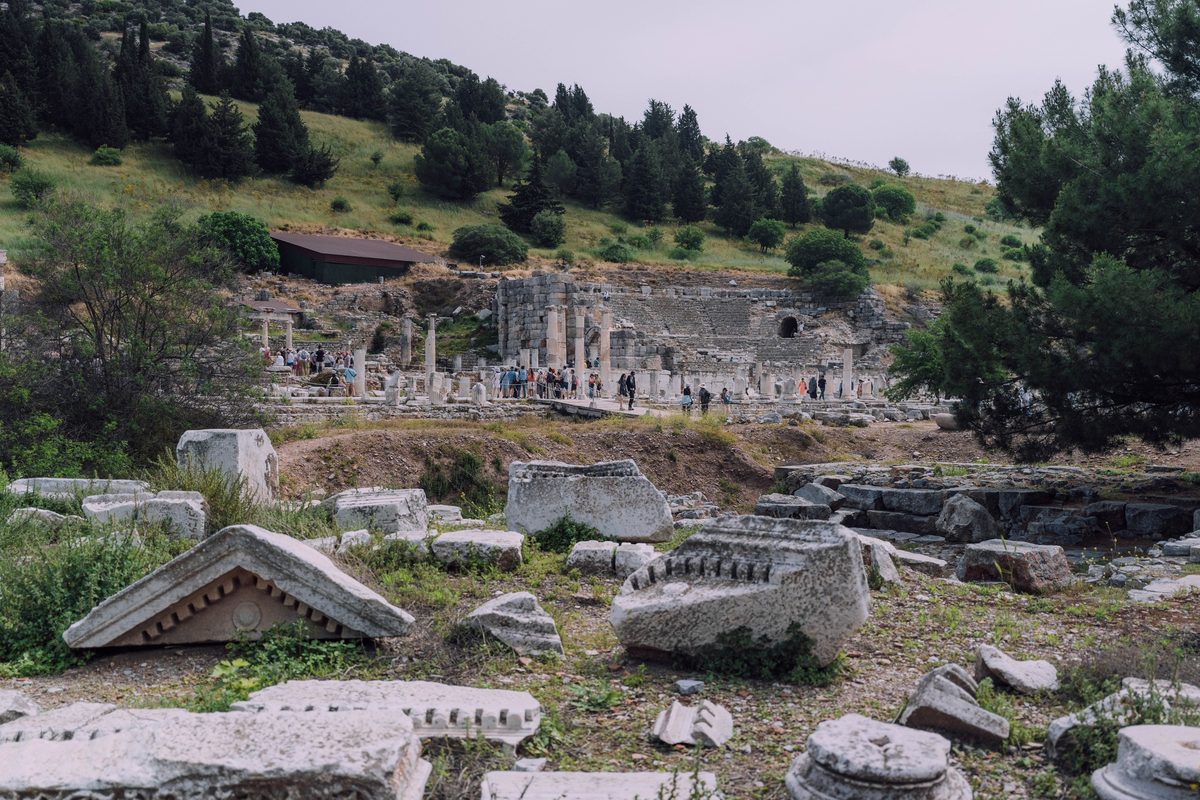 Carved marble blocks in the grass of the State Agora at Ephesus, with visitors and the seats of the Odeon on the hillside behind