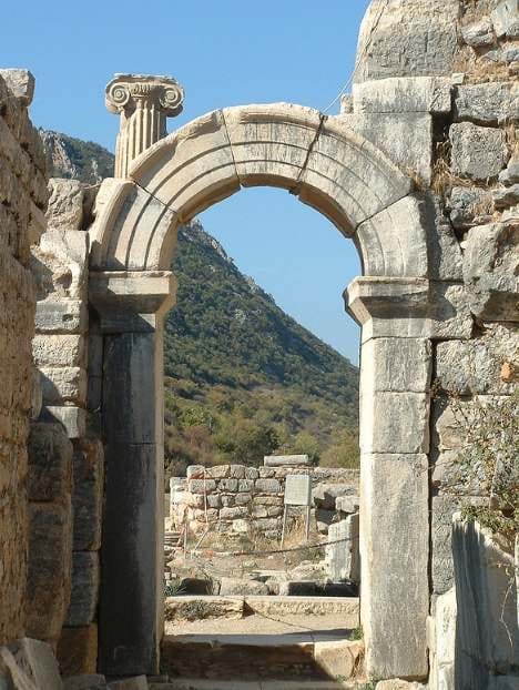 Well-preserved stone arch at Ephesus framing a green hillside, with an Ionic capital behind it