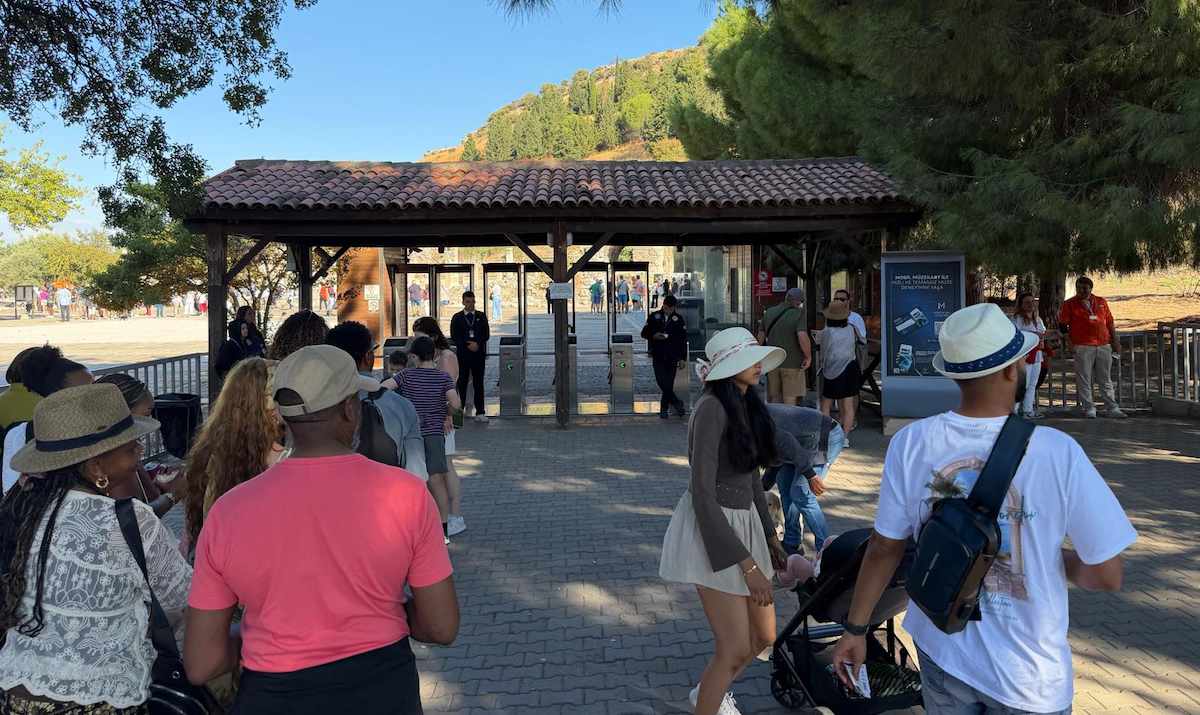 Visitors walking toward the turnstiles under a tiled-roof pavilion at the Upper Gate of Ephesus, with security staff at the entrance and a pine-covered hillside behind