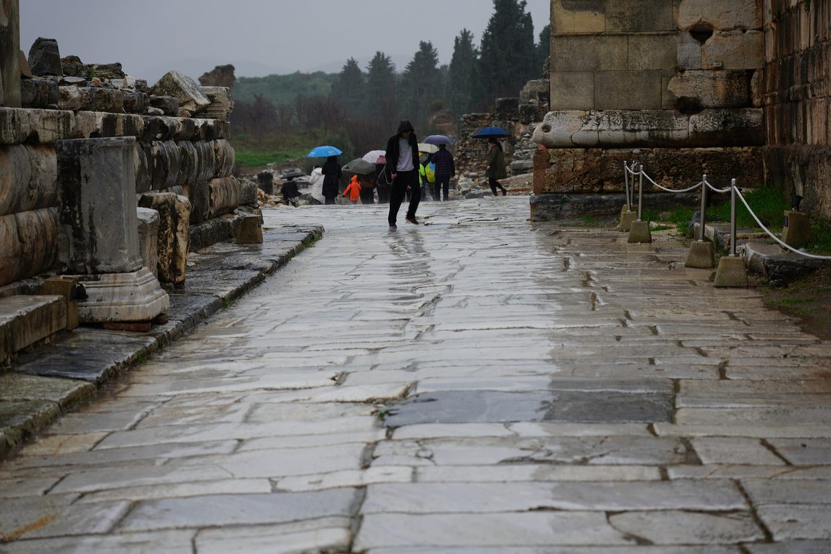 Wet marble paving stones shining on an ancient street at Ephesus on a rainy winter day, with visitors under umbrellas in the distance