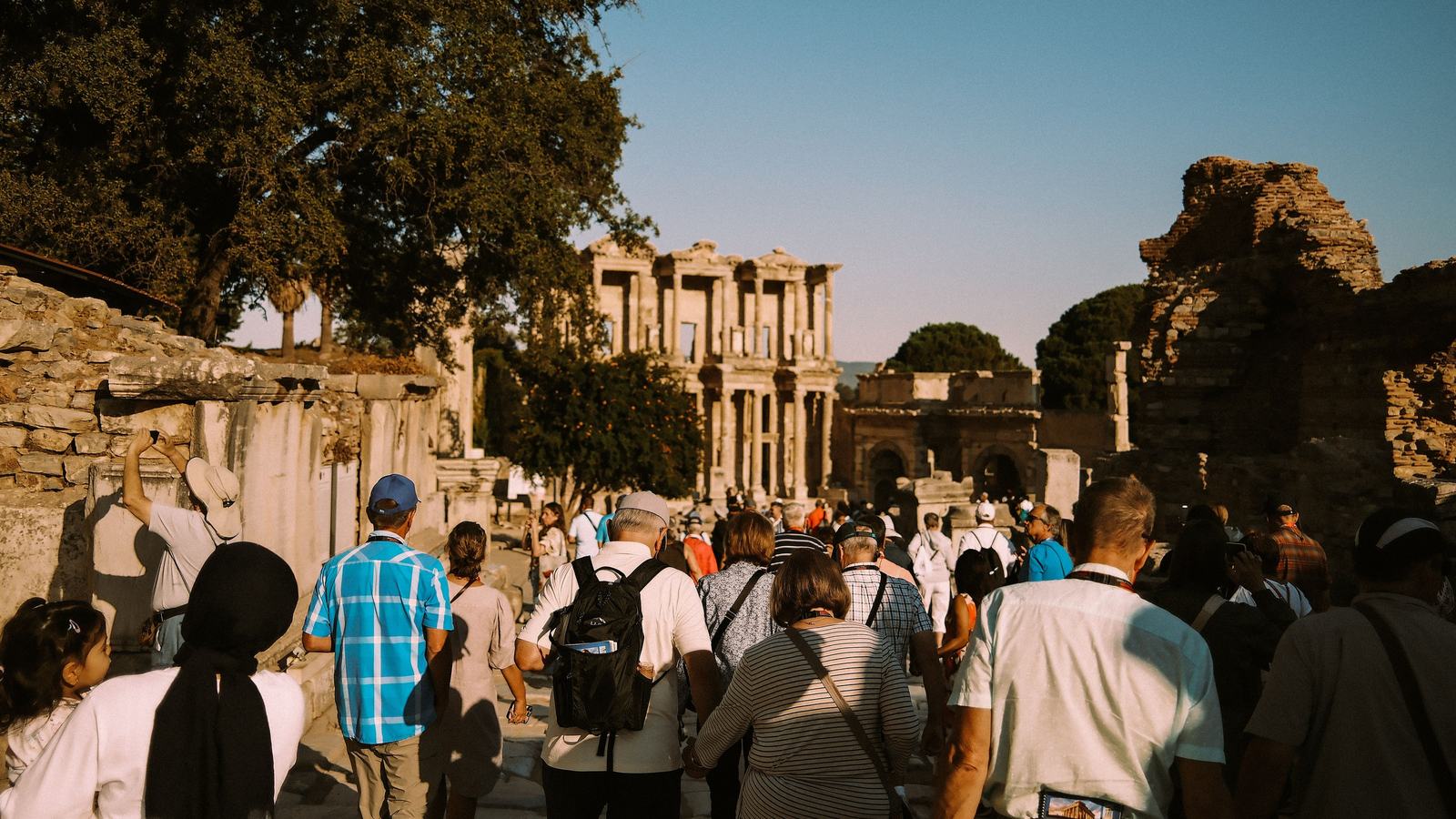 Cruise passengers with lanyards walking down Curetes Street at Ephesus toward the Library of Celsus in morning sun, seen from behind