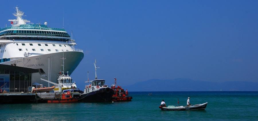 A small wooden fishing boat with two fishermen on the turquoise water of Kusadasi harbor beside a moored cruise ship