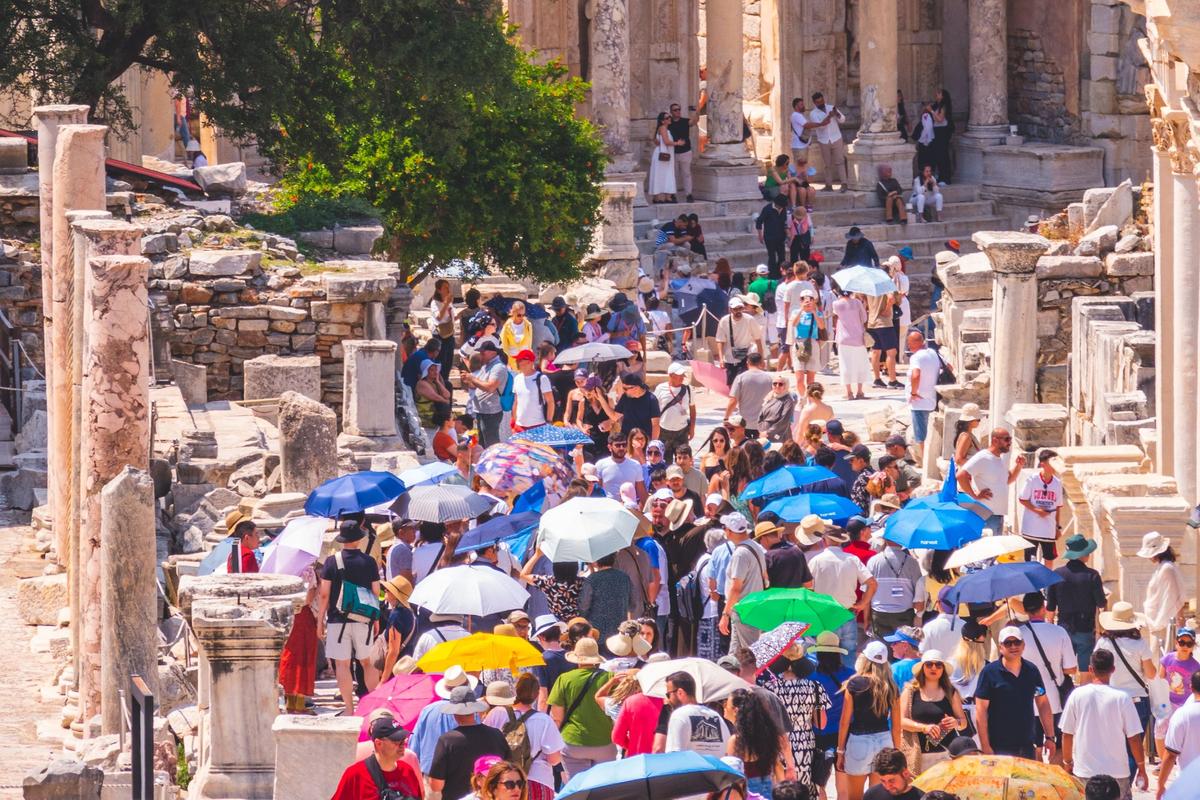 A midday summer crowd with sun hats and colorful umbrellas on the marble street below the steps and lower columns of the Library of Celsus at Ephesus