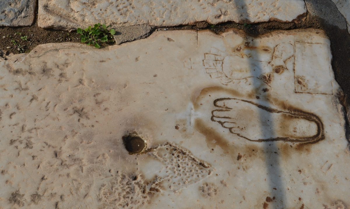 Ancient graffiti carved into a marble paving slab at Ephesus, showing a footprint and a female figure.