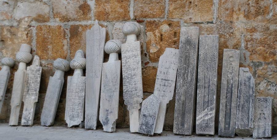 Carved Ottoman marble tombstones, several topped with stone turbans, leaning in a row against an old stone wall.