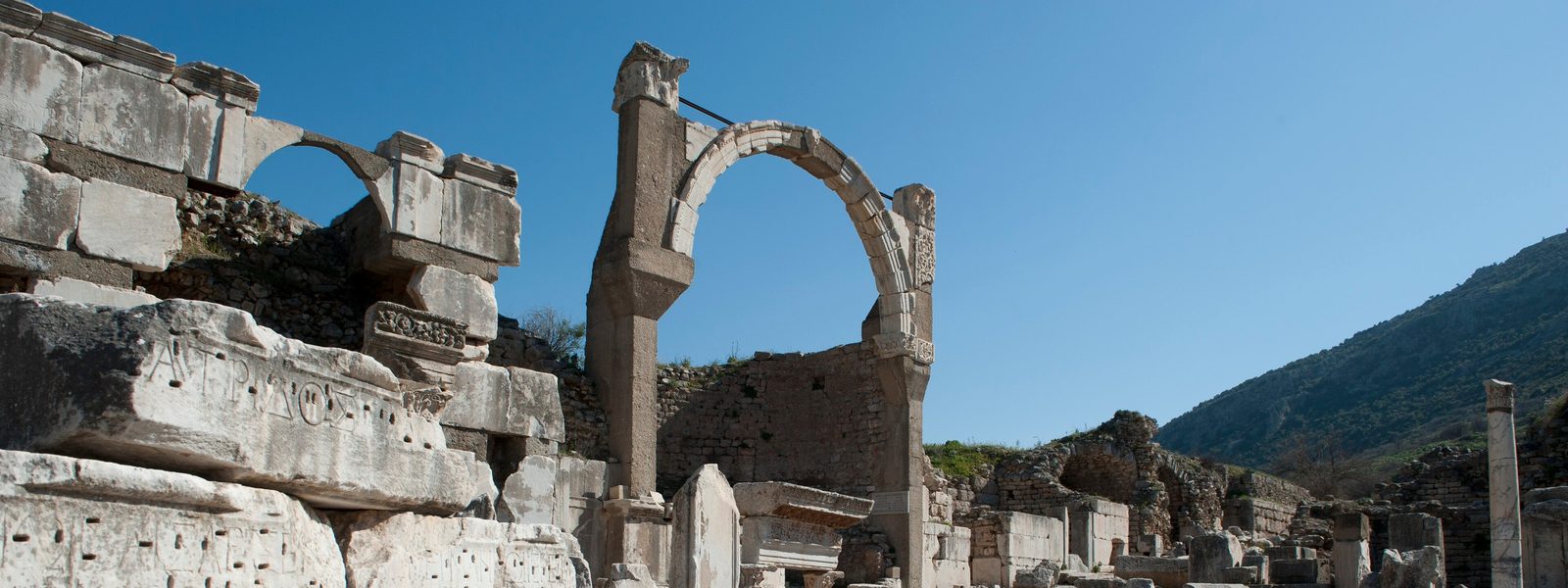 The tall arch of the Pollio Fountain at Ephesus beside inscribed marble blocks of the Pollio Monument, under a clear blue sky