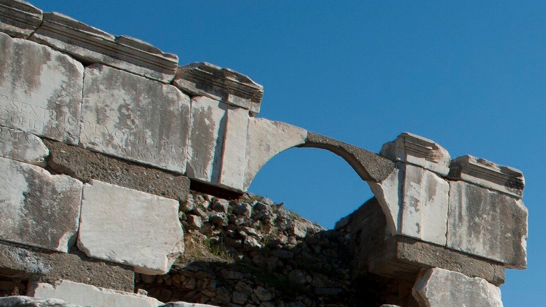 Weathered marble blocks of the Pollio Monument at Ephesus carved with a Latin inscription naming Sextilius Pollio, beside a small stone arch