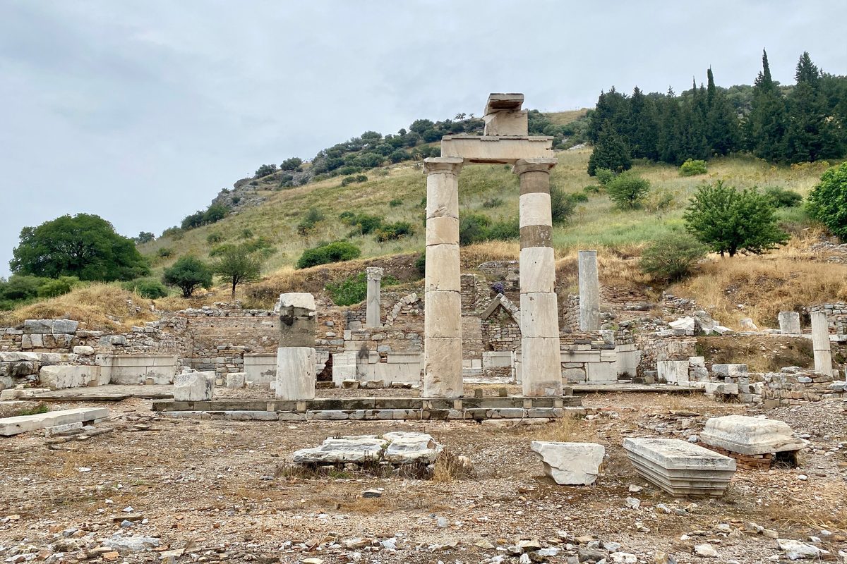 Two re-erected Doric columns of the Prytaneion at Ephesus among low ruined walls, with a green hillside and cypress trees behind