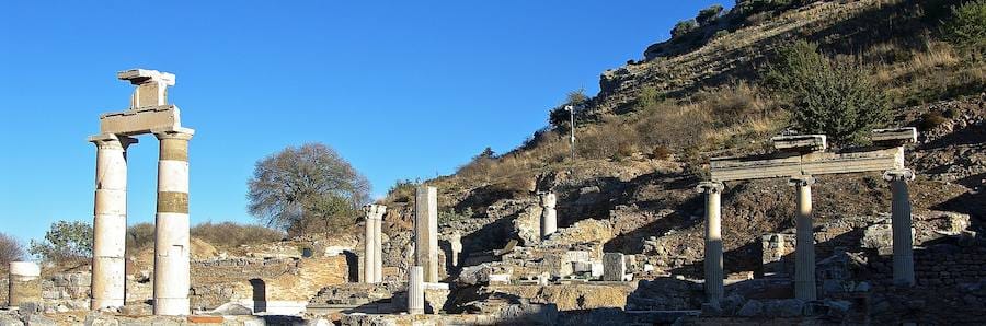 Re-erected columns and architraves of the Prytaneum at Ephesus below a dry hillside under a deep blue sky.