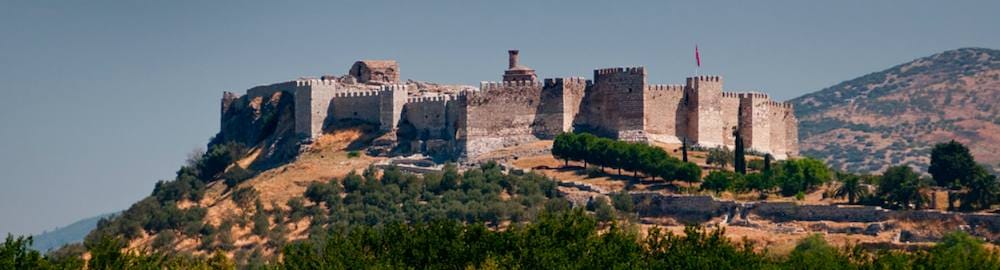 The crenellated walls and towers of Ayasuluk Fortress on a hill above Selcuk, surrounded by olive trees.