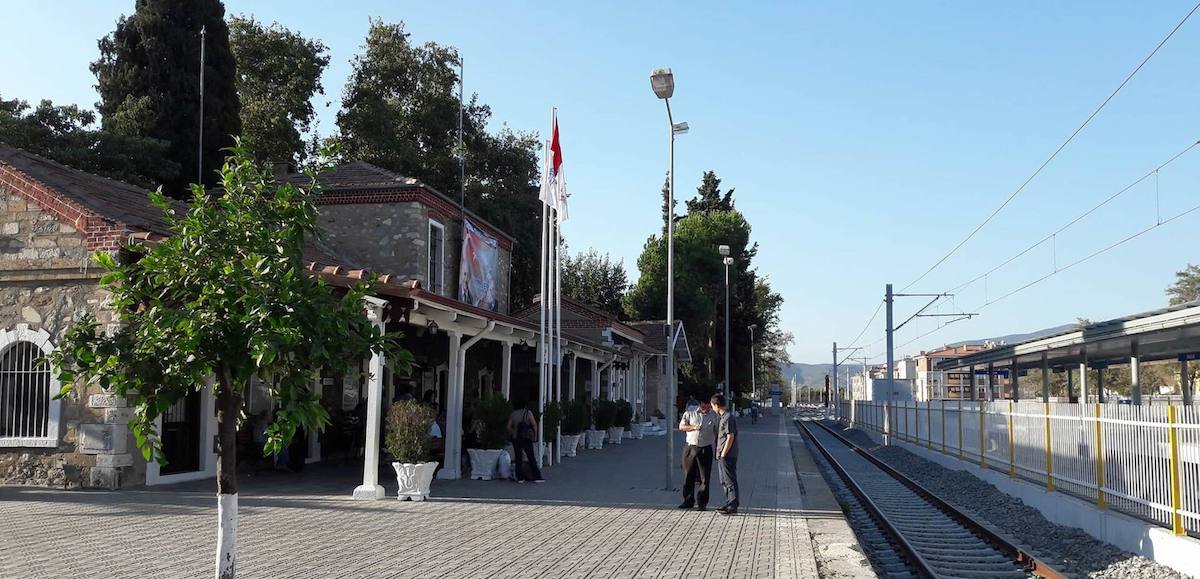 The old stone station building at Selcuk with tiled roofs, a covered porch and a Turkish flag, beside the platform and the electrified line