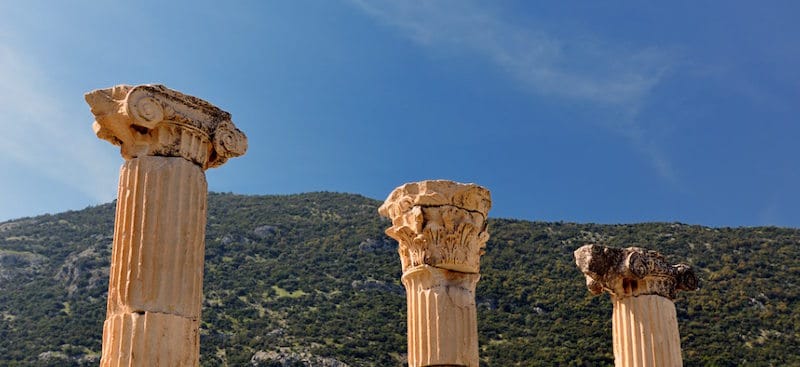 Three ancient column capitals, one Ionic and two Corinthian, rise against a deep blue sky above a green hillside.